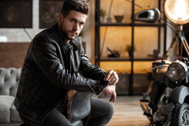 Deri ceketli yakışıklı genç adam taburede otururken gözlerini kaçırıyor.