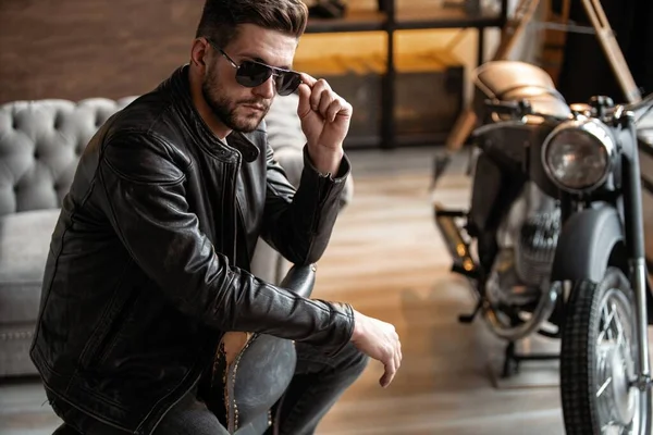Handsome young man in leather jacket looking away while sitting on the stool