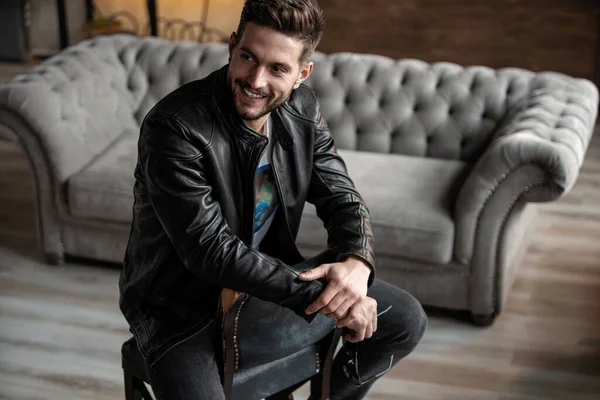 Handsome young man in leather jacket looking away while sitting on the stool — Stock Photo, Image