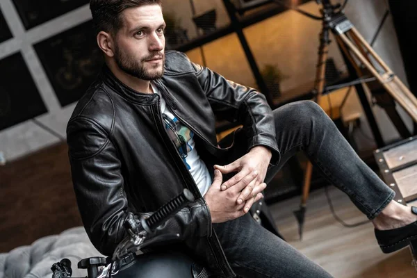 Handsome young man in leather jacket looking away while sitting on the stool — Stock Photo, Image