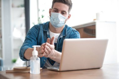 Coronavirus. Koruyucu maske takan bir adam evde çalışıyor. Ellerini dezenfektan jeliyle temizliyor.