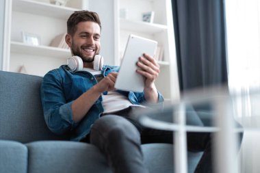 Çekici genç adam tablet kullanıyor ve evinde kanepede yatarken gülümsüyor..