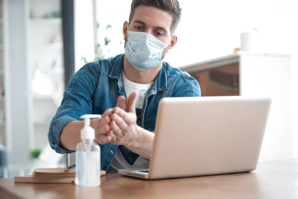 Coronavirus. Hombre trabajando desde casa usando máscara protectora. Limpiándose las manos con gel desinfectante — Foto de Stock