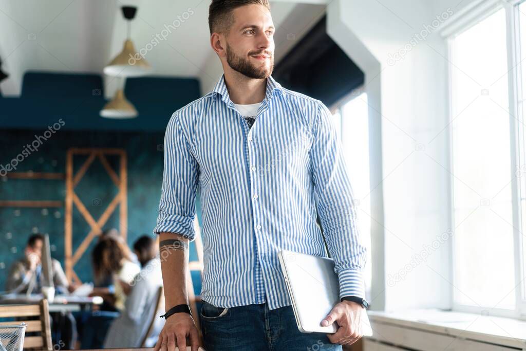 Happy businessman standing in the office with coworkers in the background working by the desk.