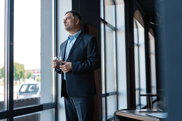 Hombre de negocios maduro bebiendo agua y mirando por una ventana a la ciudad desde un edificio de oficinas . —  Fotos de Stock