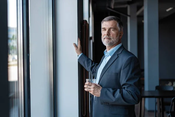 Volwassen zakenman drinkend water en kijkend uit een raam naar de stad vanuit een kantoorgebouw. — Stockfoto