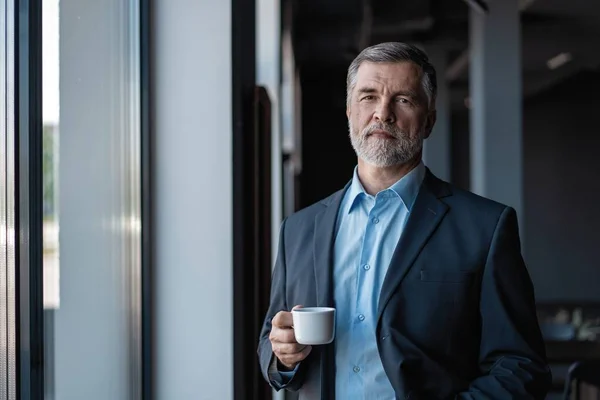 Volwassen zakenman drinkt een koffie en kijkt uit een raam naar de stad vanuit een kantoorgebouw. — Stockfoto