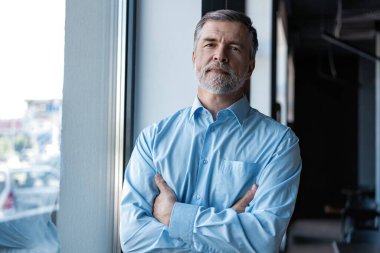 Ofis binasının penceresinin yanında dururken kollarını kavuşturup kameraya bakan neşeli iş adamı..