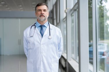 Özgüvenli gülümseyen doktor hastanede poz veriyor ve tıbbi ekip arka planda çalışıyor..