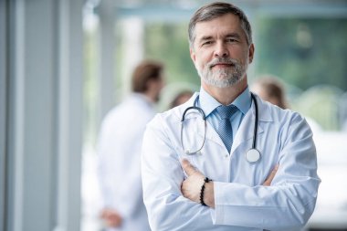 Özgüvenli gülümseyen doktor hastanede poz veriyor ve tıbbi ekip arka planda çalışıyor..