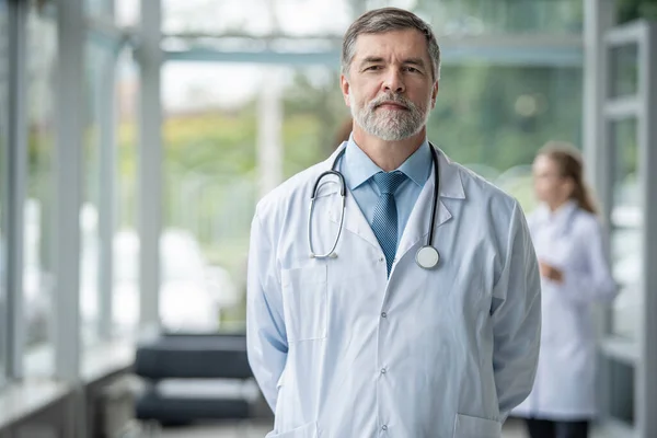 Médico sonriente confiado posando en el hospital con el equipo médico trabajando en el fondo . — Foto de Stock