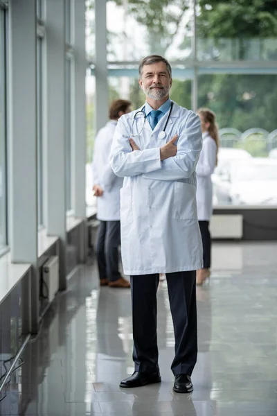 Σίγουρος χαμογελαστός γιατρός που ποζάρει στο νοσοκομείο με την ιατρική ομάδα να εργάζεται στο παρασκήνιο. — Φωτογραφία Αρχείου