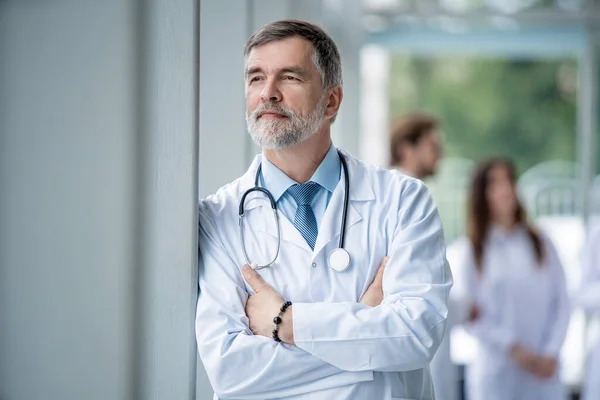 Médico sonriente confiado posando en el hospital con el equipo médico trabajando en el fondo . — Foto de Stock