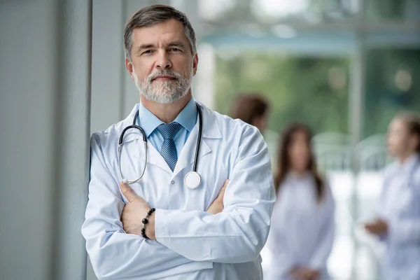 Médico sonriente confiado posando en el hospital con el equipo médico trabajando en el fondo . — Foto de Stock