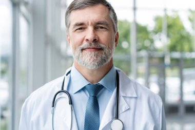 Özgüvenli gülümseyen doktor hastanede poz veriyor ve tıbbi ekip arka planda çalışıyor..