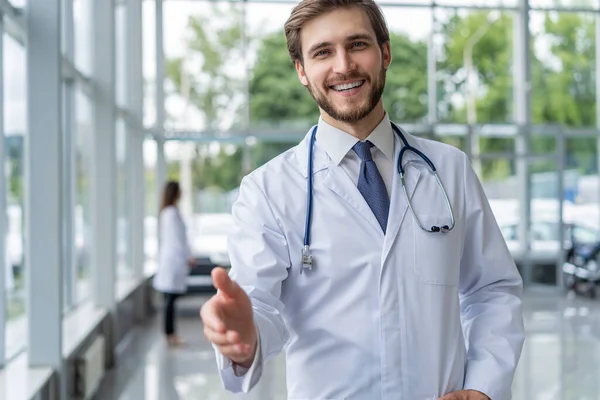 Un joven doctor dando la bienvenida en su consultorio. médico médico hombre sonrisa, toma la mano gesto de bienvenida con estetoscopio . — Foto de Stock