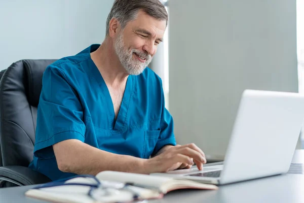 Médico varón maduro trabajando en el ordenador portátil, sentado en el consultorio médico. —  Fotos de Stock