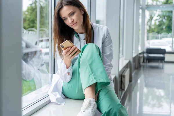 병원에서 스마트폰을 사용하면서 흰 코트를 입고 앉아 있는 젊은 여성 의사의 모습, 근무 후 긴장을 풀고 있는 모습 — 스톡 사진