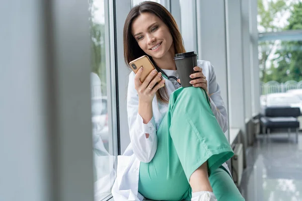병원에서 스마트폰을 사용하면서 흰 코트를 입고 앉아 있는 젊은 여성 의사의 모습, 근무 후 긴장을 풀고 있는 모습 — 스톡 사진