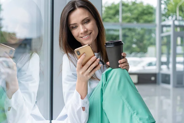 Portrait de jeune femme médecin en manteau blanc assis tout en utilisant un smartphone à l'hôpital, se détendre après la journée de travail — Photo