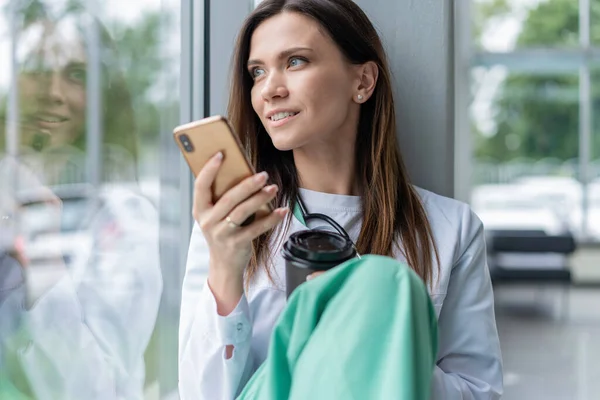 Retrato de la joven doctora en bata blanca sentada mientras usa el teléfono inteligente en el hospital, relájese después del día laboral — Foto de Stock