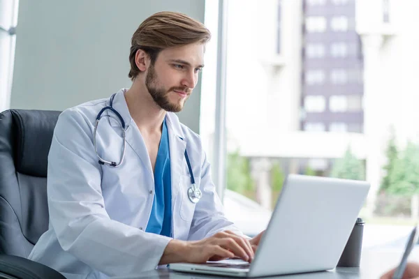 젊은 남자 의사가 노트북 컴퓨터를 사용하며 의료 실에 앉아 있는 모습. — 스톡 사진