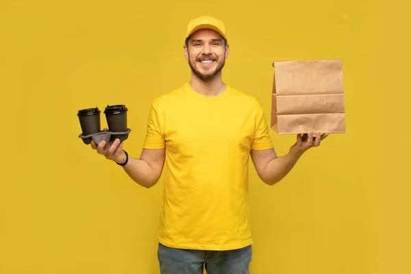 Porträt eines jungen Zustellers in gelber Uniform mit Papierschachtel und Kaffee zum Mitnehmen isoliert vor gelbem Hintergrund. — Stockfoto