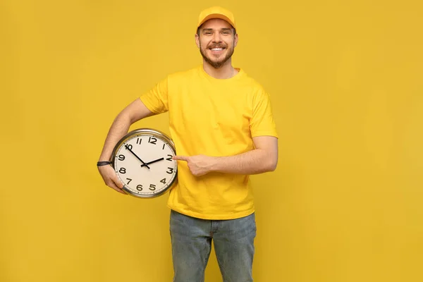 Entrega homem em amarelo workwear segurar relógio isolado no fundo amarelo estúdio retrato . — Fotografia de Stock