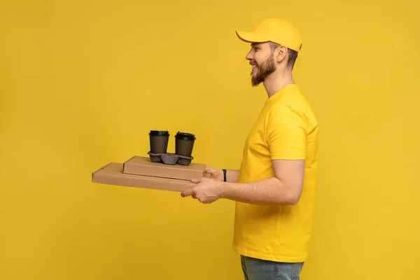 Porträt eines jungen Zustellers in gelber Uniform mit Pizzakartons und Kaffee zum Mitnehmen isoliert vor gelbem Hintergrund. — Stockfoto