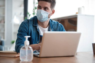 Evden çalışan genç beyaz bir adam koruyucu maske takıyor, dizüstü bilgisayar kullanıyor. Coronavirus salgını, covid 19 karantina