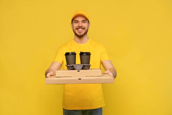Porträt eines jungen Zustellers in gelber Uniform mit Pizzakartons und Kaffee zum Mitnehmen isoliert vor gelbem Hintergrund. — Stockfoto