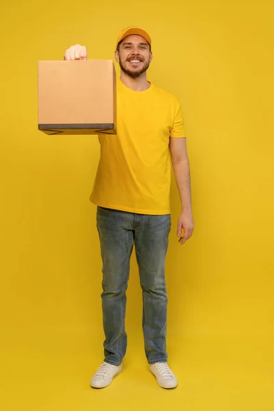 Portrait of excited delivery man in yellow uniform holding paper box isolated over yellow background. — Stock Photo, Image