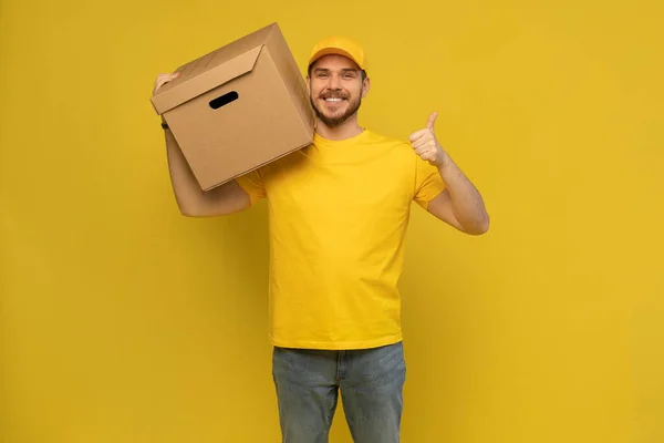 Portrait of excited delivery man in yellow uniform holding paper box isolated over yellow background. — Stock Photo, Image