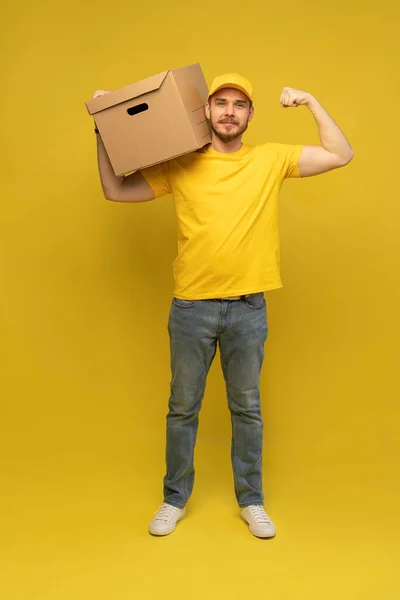 Portrait of excited delivery man in yellow uniform holding paper box isolated over yellow background. — Stock Photo, Image