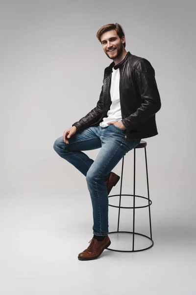 Moda adamı, yakışıklı, ciddi, güzel erkek manken portresi deri ceket giy, beyaz arka planda sandalyede otur.. — Stok fotoğraf