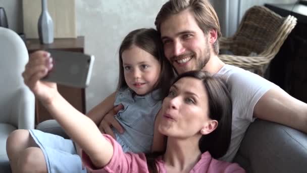 Família feliz sentada no sofá, mãe sorridente segurando celular, tirando selfie. casal animado posando para foto com filha — Vídeo de Stock