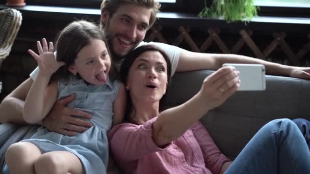 Familia feliz sentada en el sofá, madre sonriente sosteniendo el teléfono móvil, tomando selfie. Emocionada pareja posando para la foto con su hija — Vídeos de Stock