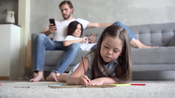 Счастливое семейное времяпрепровождение дома, пара родителей расслабляется, разговаривая на диване в уютной гостиной, освещенной светом, в то время как маленькая дочь наслаждается активностью, играя в рисование на теплом полу. — стоковое видео