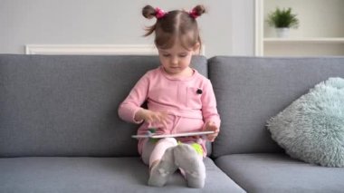 Tablet kullanan meraklı tatlı bir kız. Kanepede tek başına yatmak. Küçük çocuk evde komik oyunlar oynuyor.