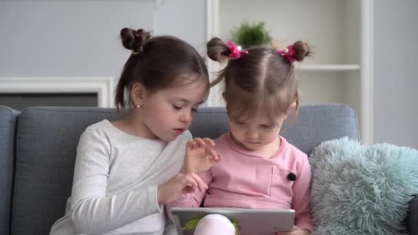 Hermanitas lindas sentadas una cerca de la otra, viendo algo en el dispositivo de la tableta o jugando un juego en él. — Vídeo de stock