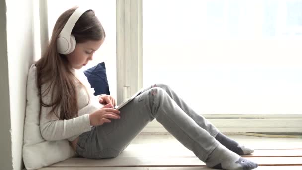 Süßes kleines Mädchen, auf einem Tablet spielend, auf einem Fensterschild sitzend. — Stockvideo