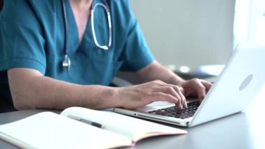 Nitelikli erkek terapist online araştırma yapıyor, haberleri okuyor, tıp dergisine notlar yazıyor. Ofis masasında dizüstü bilgisayar kullanan yaşlı doktor.