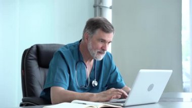 Nitelikli erkek terapist online araştırma yapıyor, haberleri okuyor, tıp dergisine notlar yazıyor. Ofis masasında dizüstü bilgisayar kullanan yaşlı doktor.