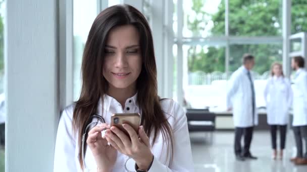 Mooie vrouwelijke arts in lab jas staan in kliniek en scrollen op smartphone scherm. — Stockvideo