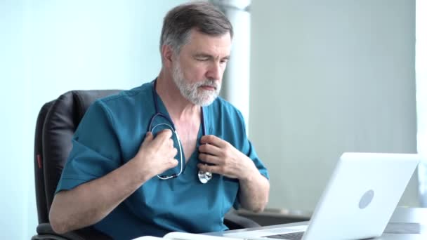Квалифицированный серьезный мужчина-терапевт проводит онлайн-исследования, читает новости, делает заметки в медицинском журнале. Сосредоточенный старый врач, использующий ноутбук в офисе — стоковое видео