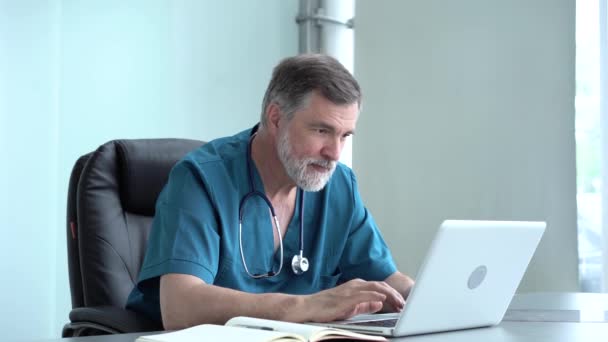 Квалифицированный серьезный мужчина-терапевт проводит онлайн-исследования, читает новости, делает заметки в медицинском журнале. Сосредоточенный старый врач, использующий ноутбук в офисе — стоковое видео