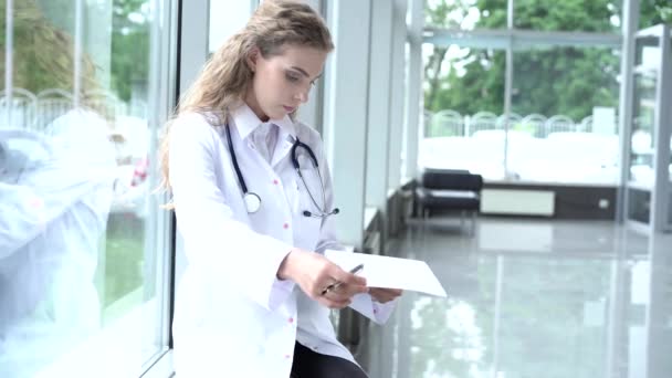 Médico sonriente con una carpeta en uniforme sentado. Atractivo médico en bata blanca y estetoscopio sonriendo a la cámara en la clínica — Vídeo de stock