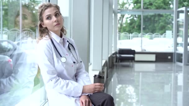 Belle femme médecin au bureau, elle prend une pause, boit du café et très fatiguée en regardant par la fenêtre panoramique. — Video