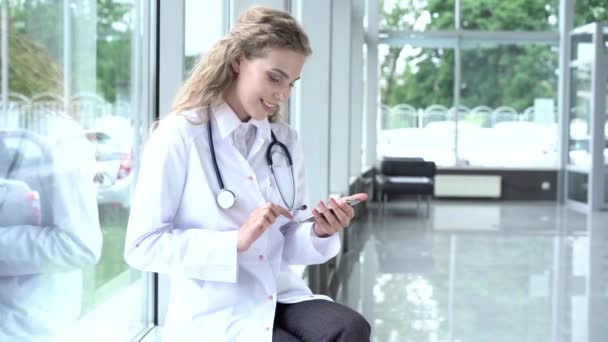 Mooie vrouwelijke arts in lab jas zitten in kliniek en scrollen op smartphone scherm. — Stockvideo