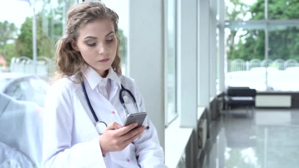 Mooie vrouwelijke arts in lab jas zitten in kliniek en scrollen op smartphone scherm. — Stockvideo